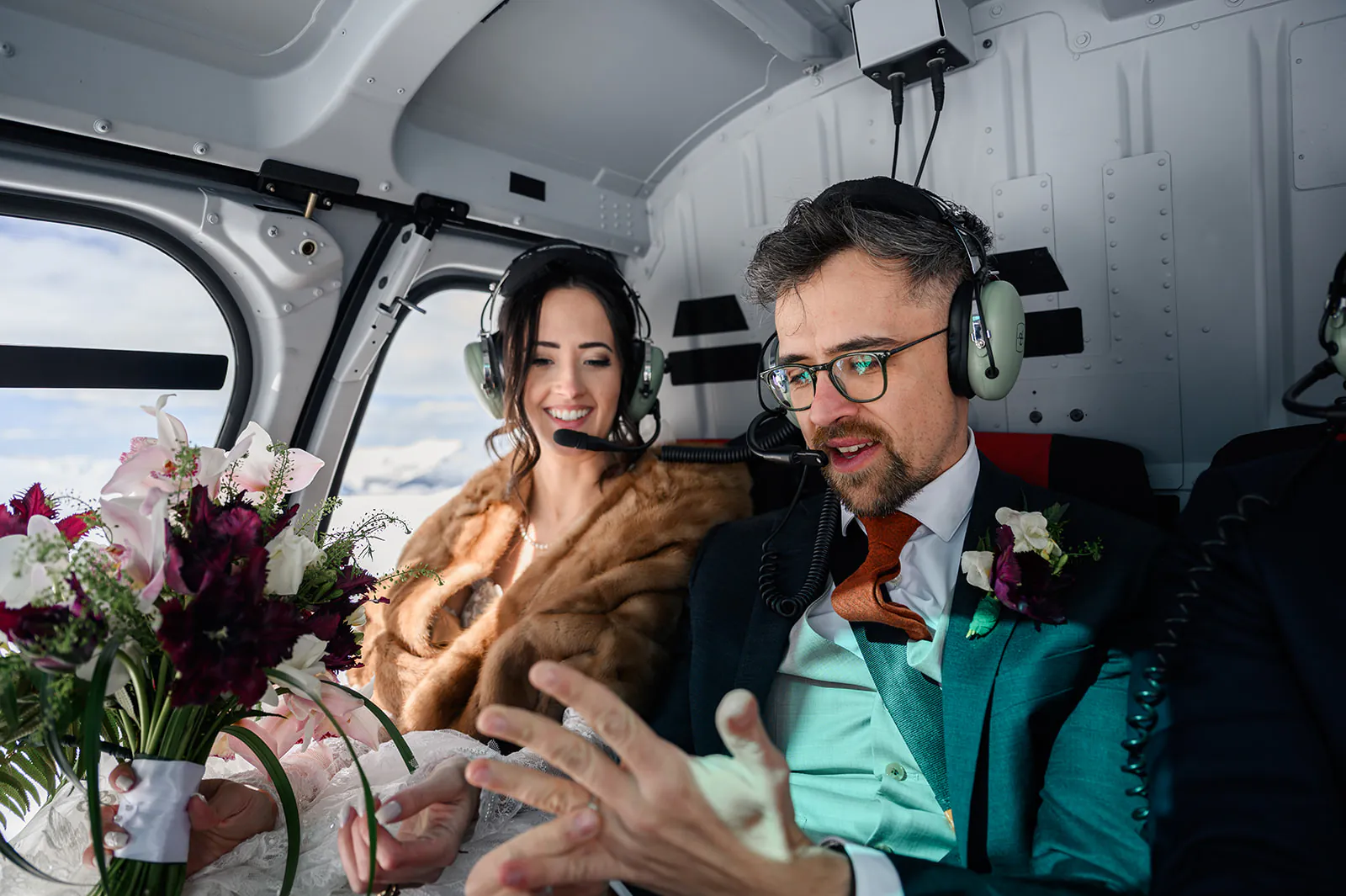 A bride and groom wearing headsets sit inside a helicopter; the groom gestures with his hands while the bride smiles, holding a bouquet of flowers.