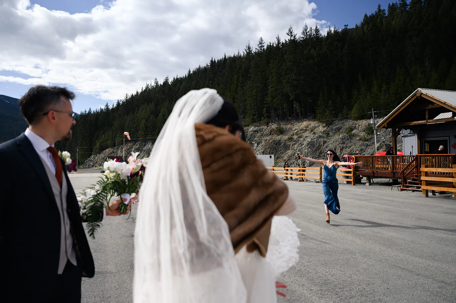 A bride in a veil and fur shawl stands next to a man in a suit as a woman in a blue dress runs toward them in an outdoor mountain setting.
