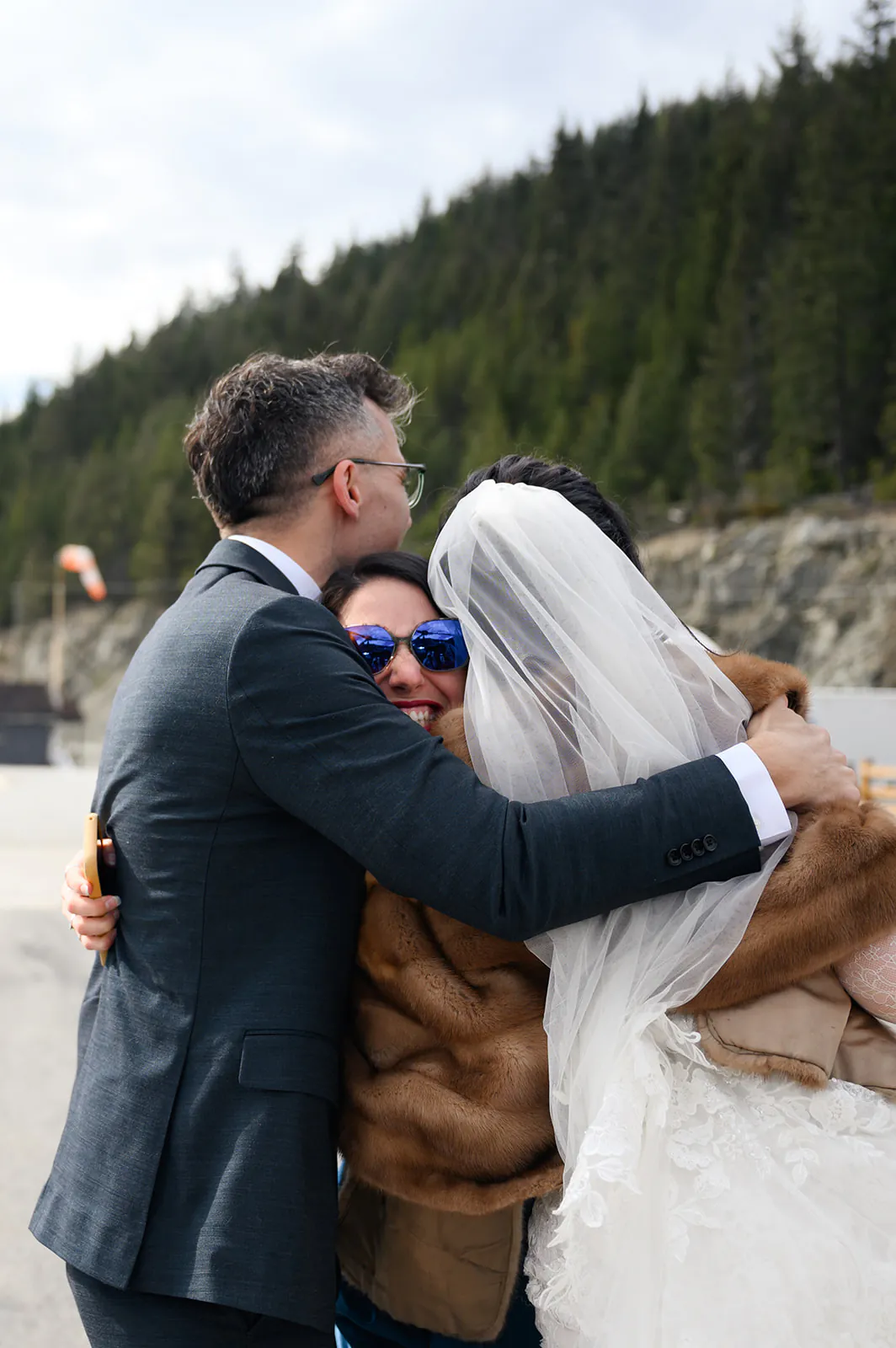A bride in a veil and a groom in a suit embrace a smiling woman wearing sunglasses outdoors near a forested hillside.