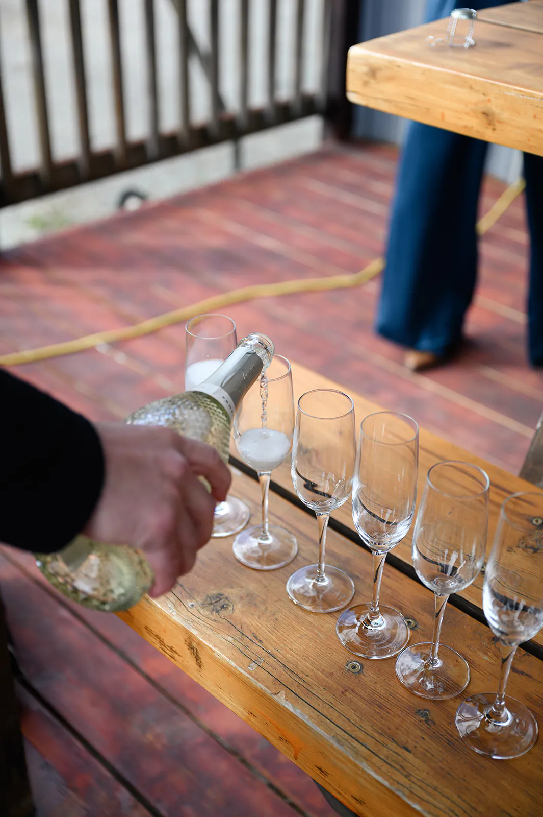 A person pours sparkling wine into a row of empty champagne flutes on a wooden table, with another person standing nearby.