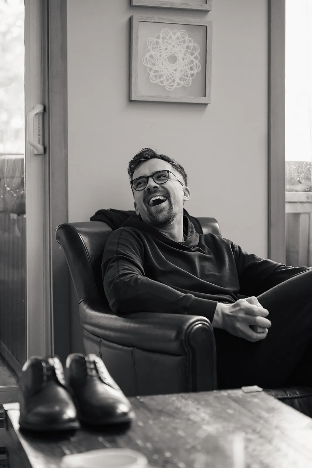 A man wearing glasses and casual clothes sits in a leather armchair, smiling and laughing. Dress shoes are visible on a nearby table. The setting appears to be indoors.