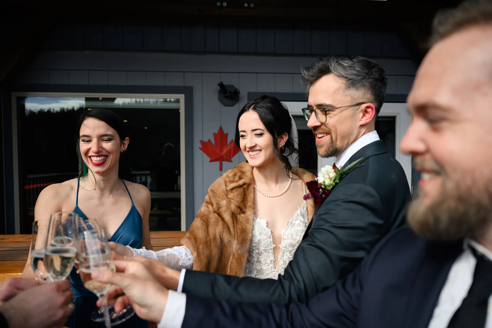 A group of people in formal attire clink glasses and smile outdoors, with a red maple leaf decoration on the building behind them.