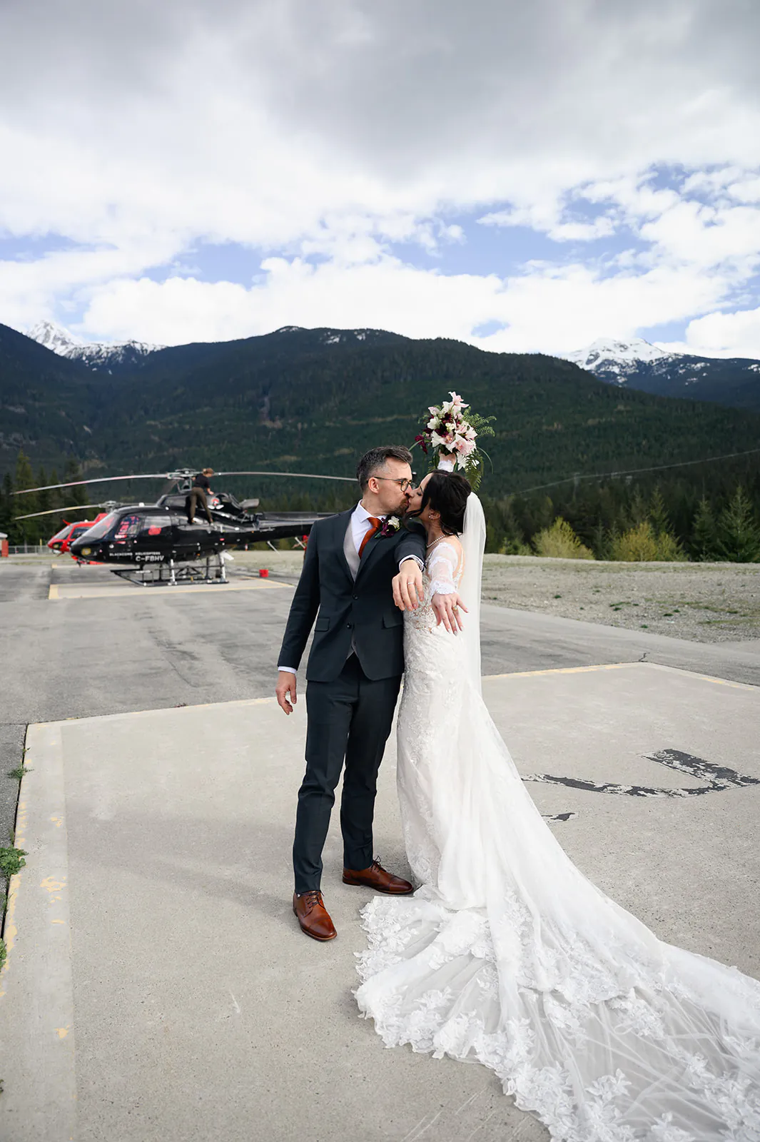 A bride and groom stand on a helipad embracing, with helicopters and forested mountains visible in the background.