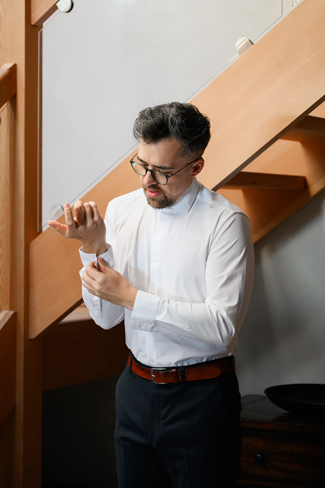 Man in glasses and a white dress shirt stands indoors by a wooden staircase, fastening the cuff of his shirt sleeve.