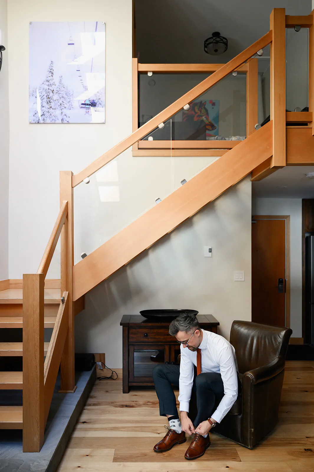 A man in formal attire sits on a chair, tying his brown shoes in a modern interior with wooden stairs and light-colored walls.