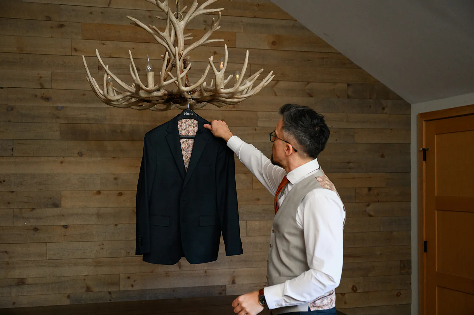 A man in a vest and tie hangs a dark suit jacket on an antler-shaped wall rack in a room with wooden walls.