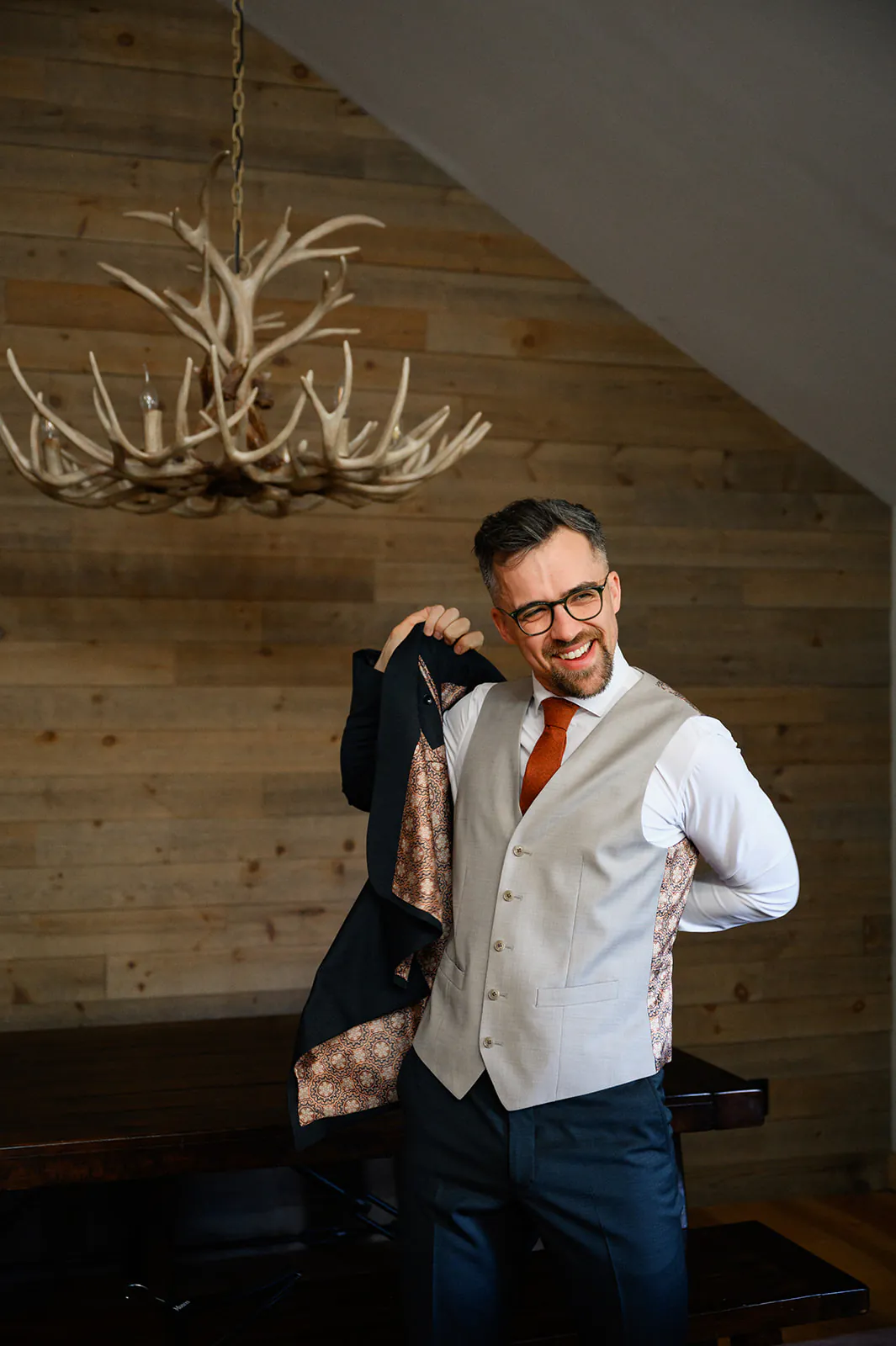 A man wearing glasses and a vest puts on a suit jacket in a room with wooden walls and an antler chandelier.