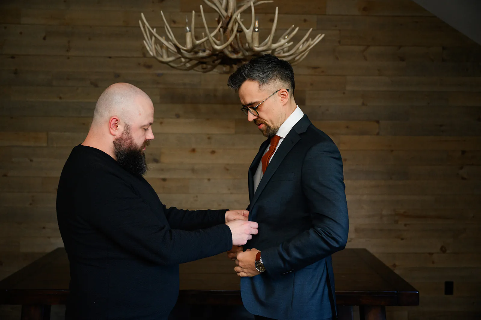 A man in casual clothes helps another man in a suit fasten his jacket in a room with wooden walls and an antler chandelier.
