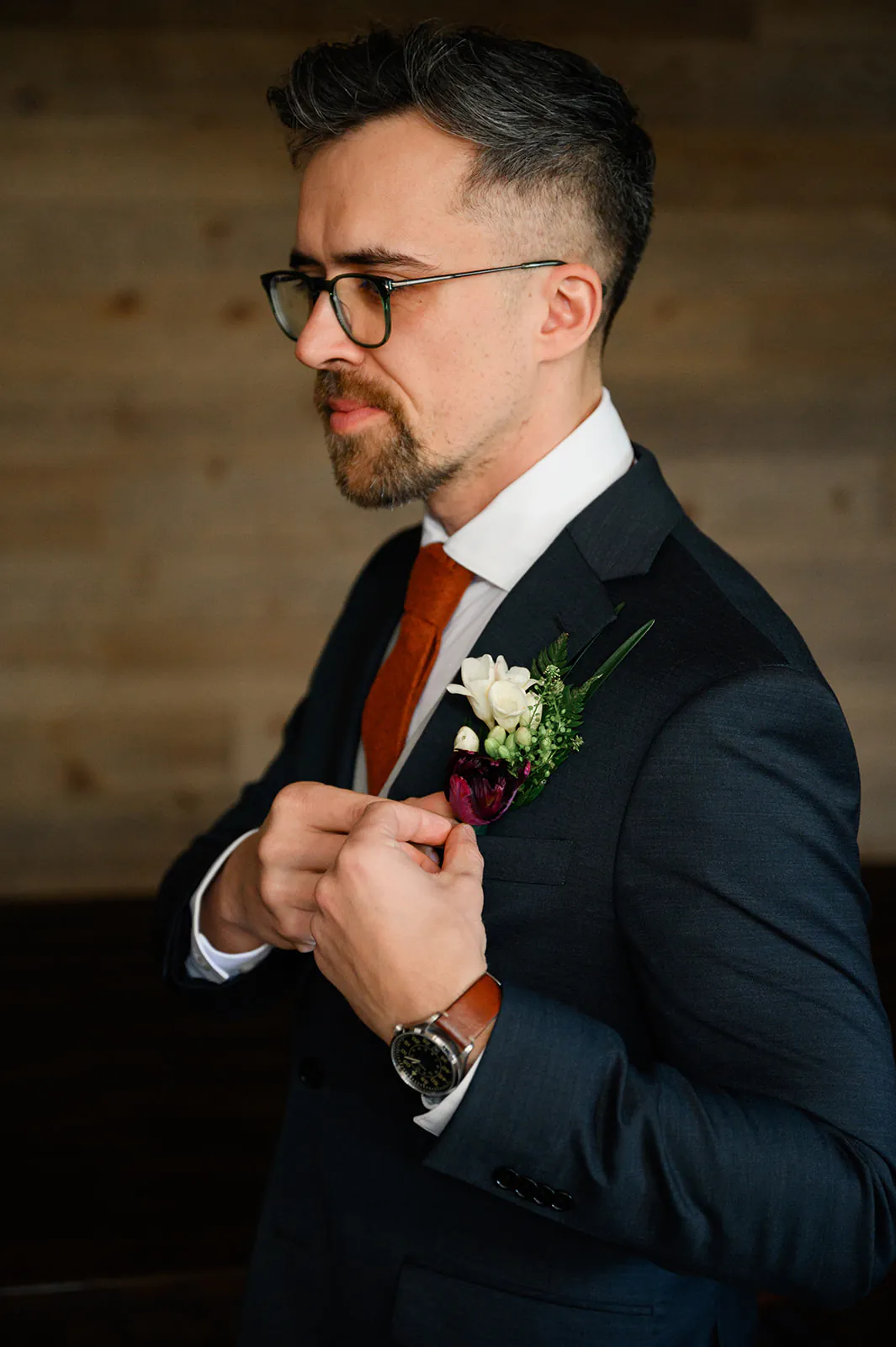 A man in a dark suit, white shirt, and orange tie adjusts his boutonniere while wearing glasses and a wristwatch, standing against a wooden background.