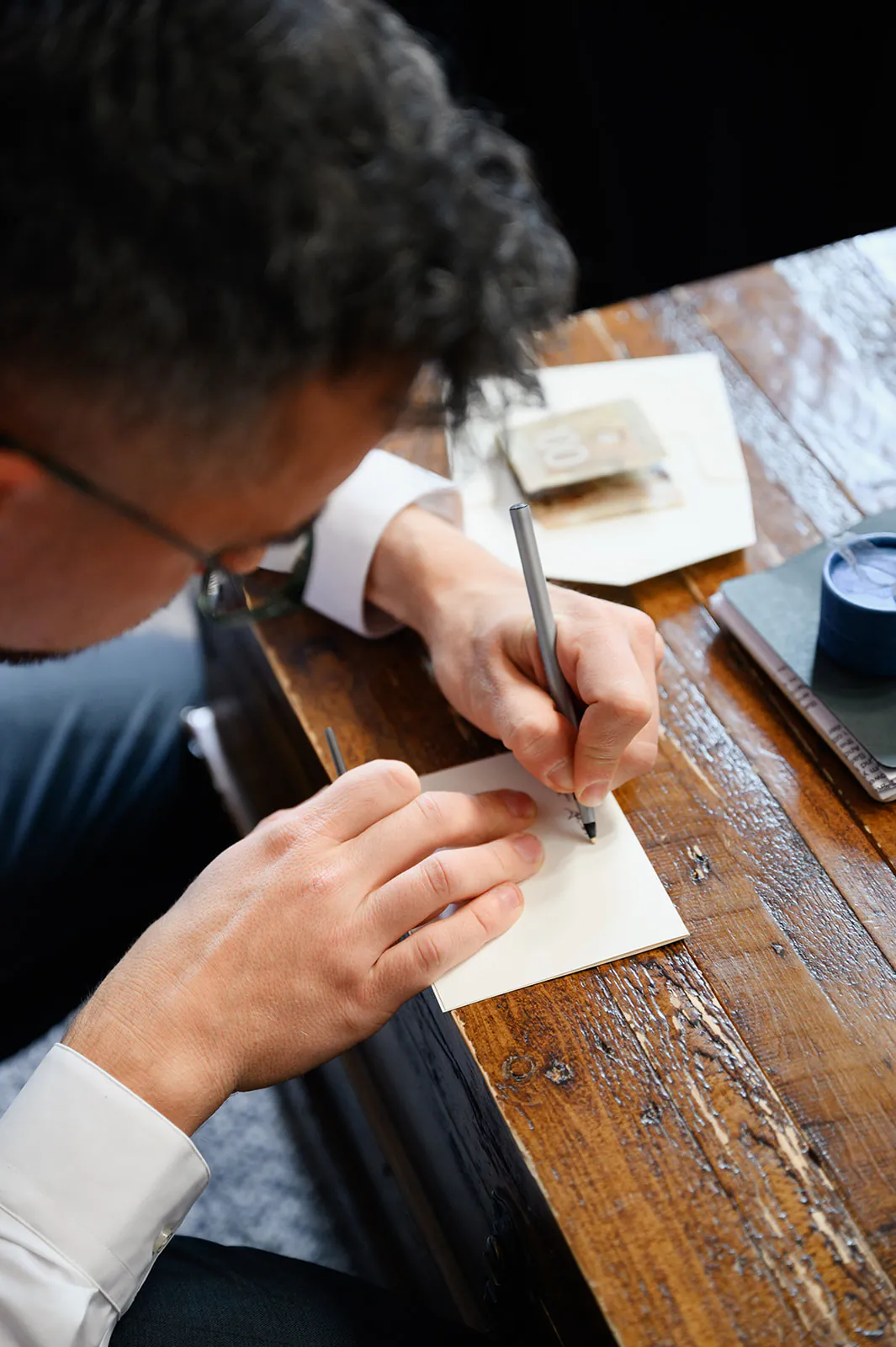 A person in a white shirt writes on a small piece of paper with a pen at a wooden table.