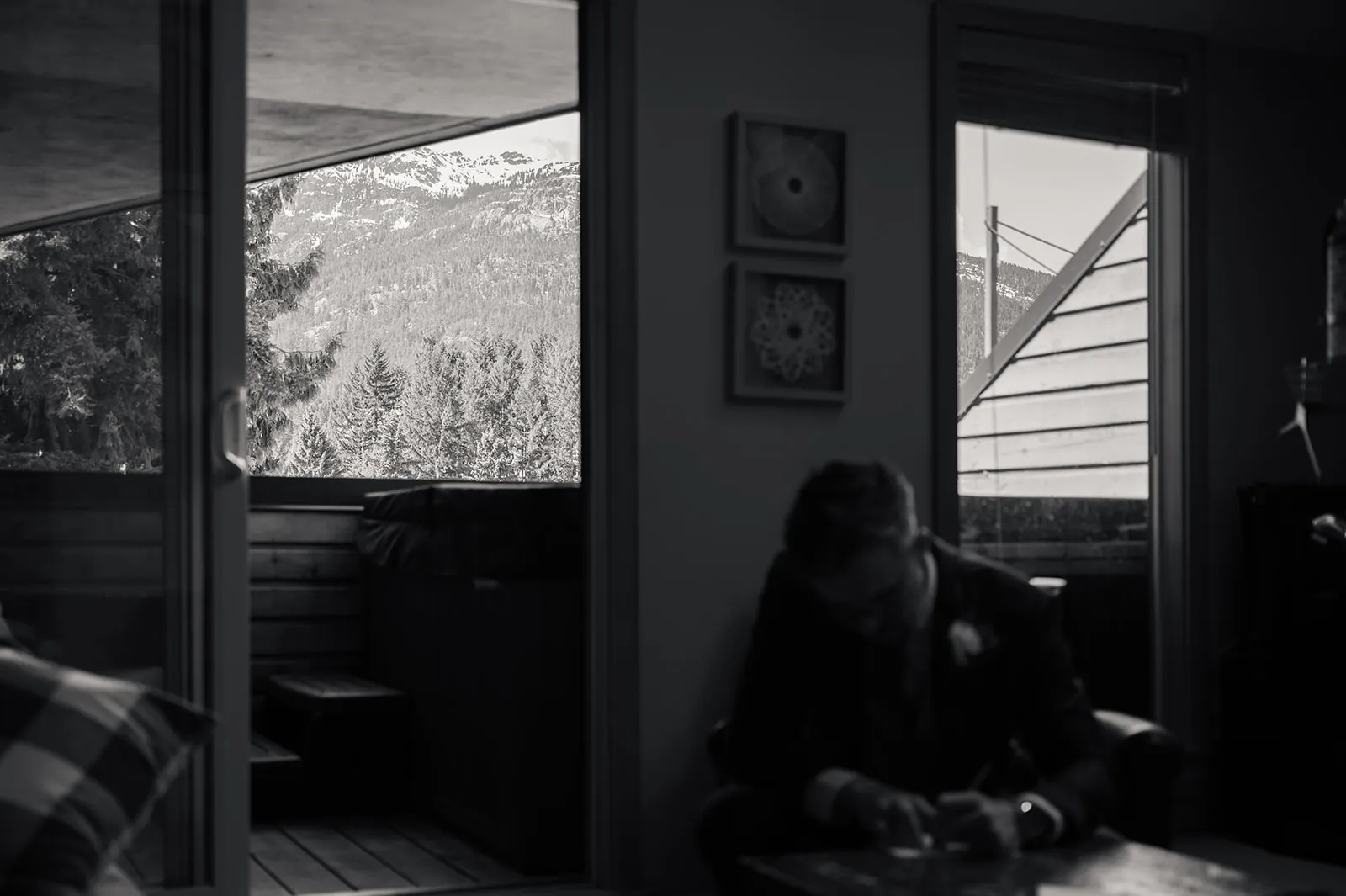 A person sits indoors writing at a table, while a balcony with trees and snow-covered mountains is visible through glass doors in the background.