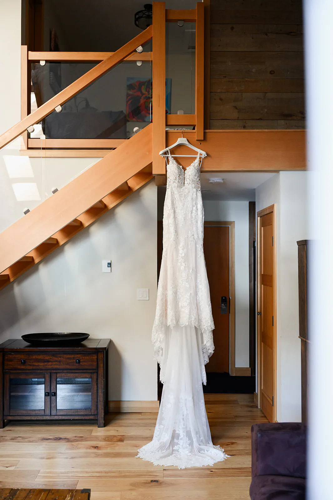A white lace wedding dress hangs on a wooden hanger from a staircase railing in a modern, sunlit interior.