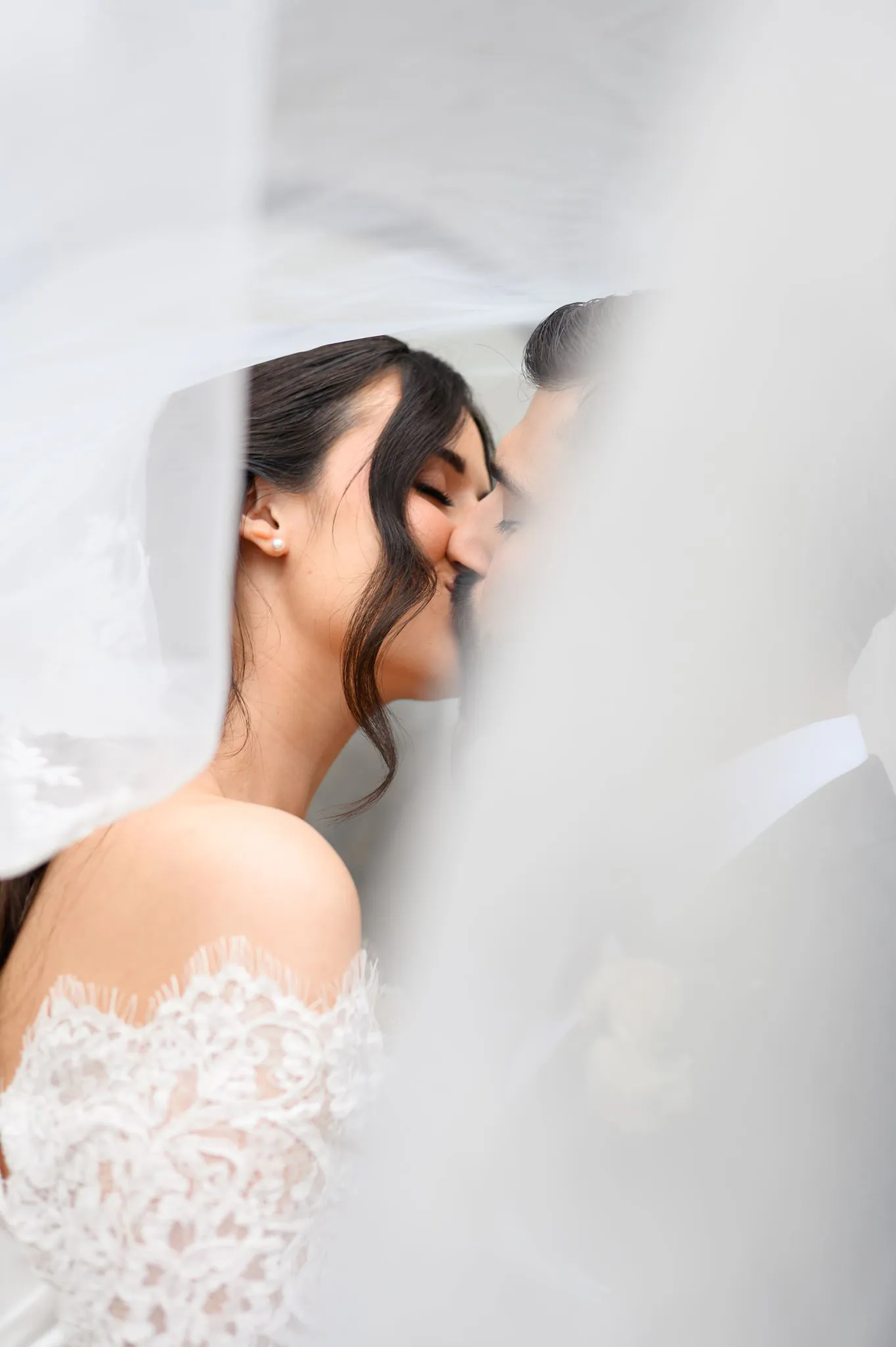 A bride and groom kiss under a sheer white veil, partially obscured, with the bride in an off-shoulder lace dress, capturing a magical wedding photography experience.