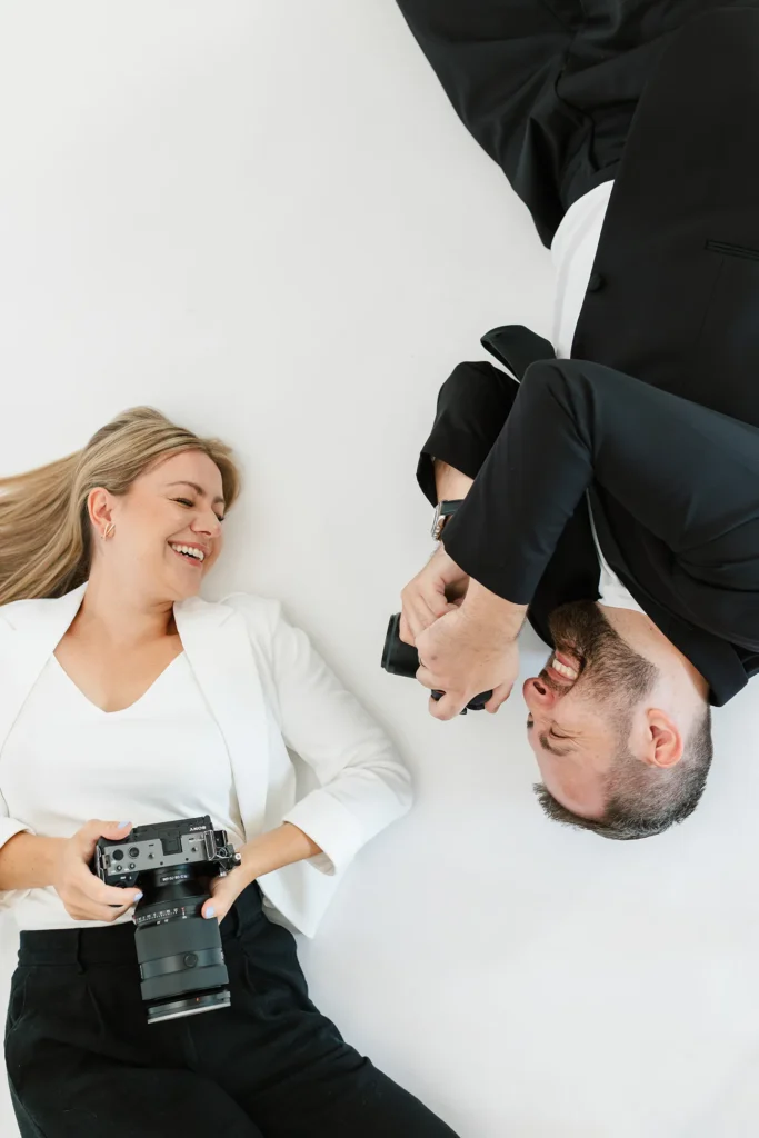 A woman and a man lie on a white surface, each holding a camera; the man points his camera at the woman while she smiles, perfectly capturing the playful spirit often found in wedding photography and videography.