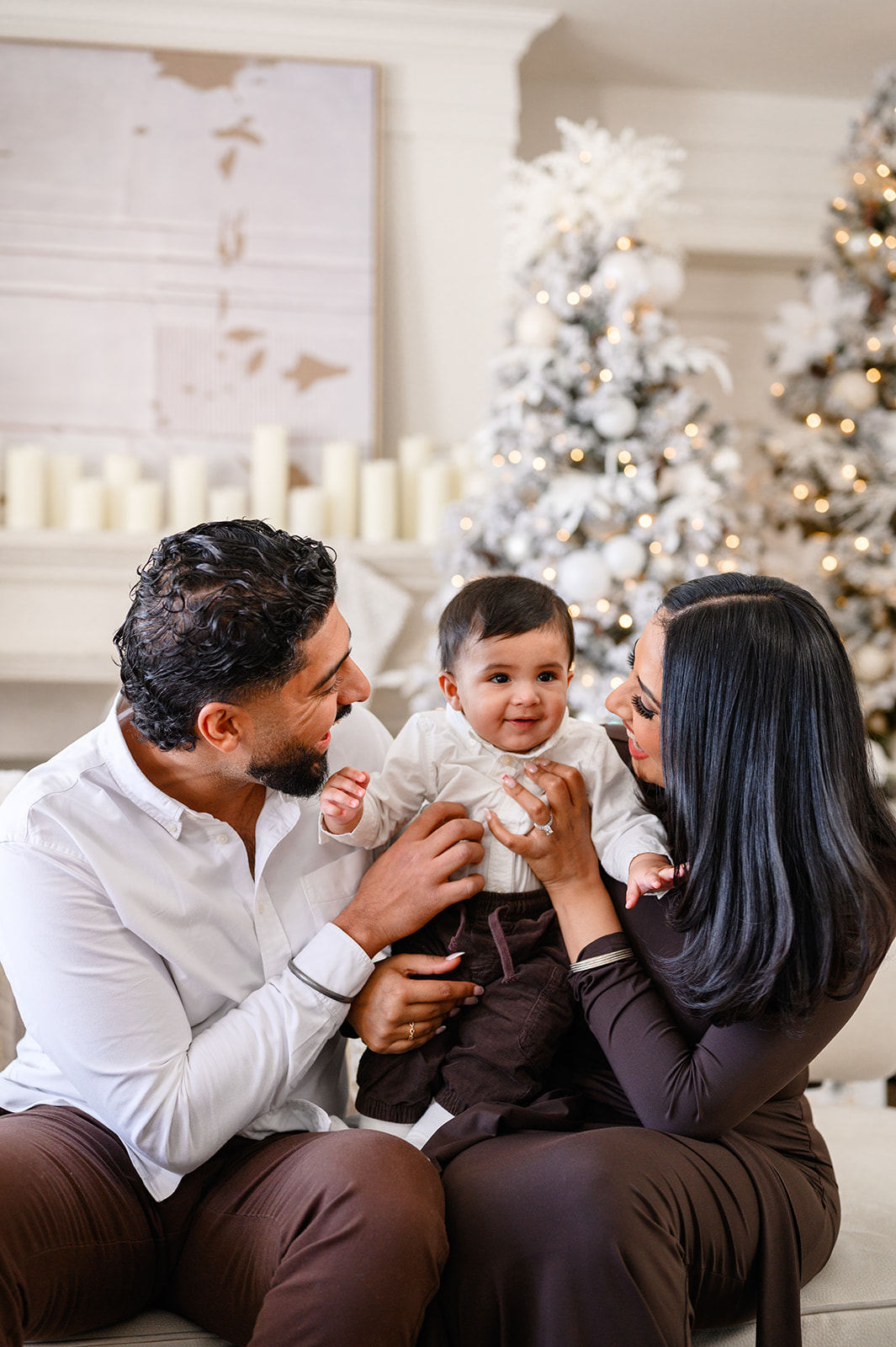 A man and woman sit on a couch holding a baby, surrounded by decorated Christmas trees and candles in the background, capturing the warmth of festive christmas minis.