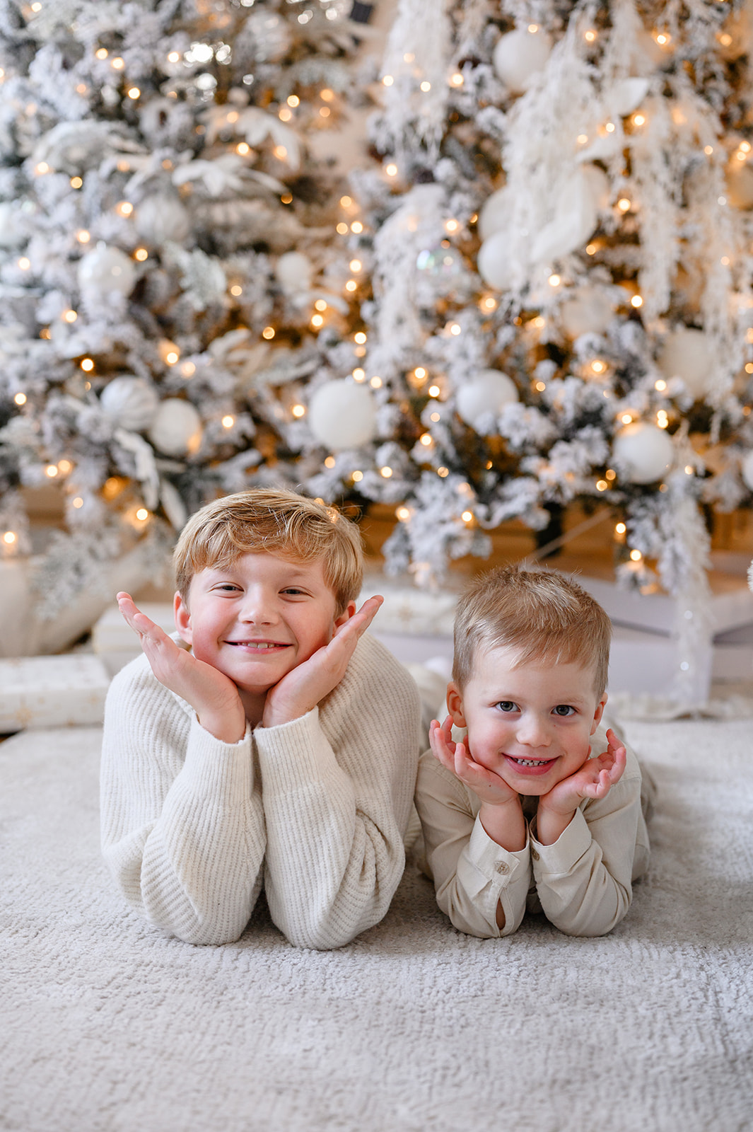 Two young boys lie on a white rug, smiling with their chins in their hands, in front of decorated Christmas trees with lights and ornaments—capturing the perfect moment for festive Christmas minis.