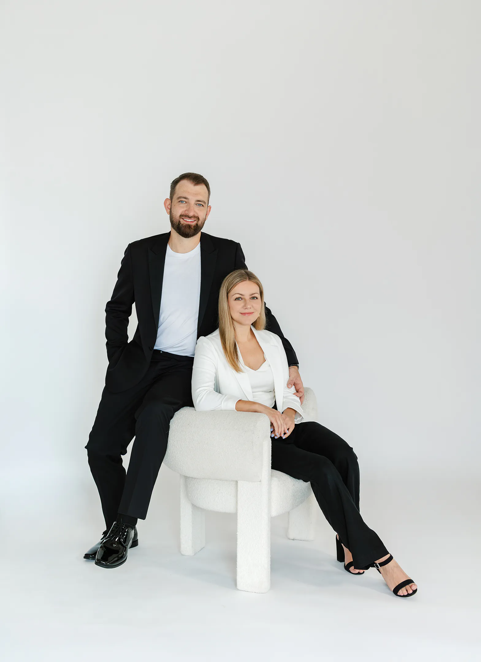 A man in a black suit stands beside a woman in a white blazer seated on a white chair, both facing the camera against a plain white background, their poised elegance reminiscent of portraits by top Toronto wedding photographers.